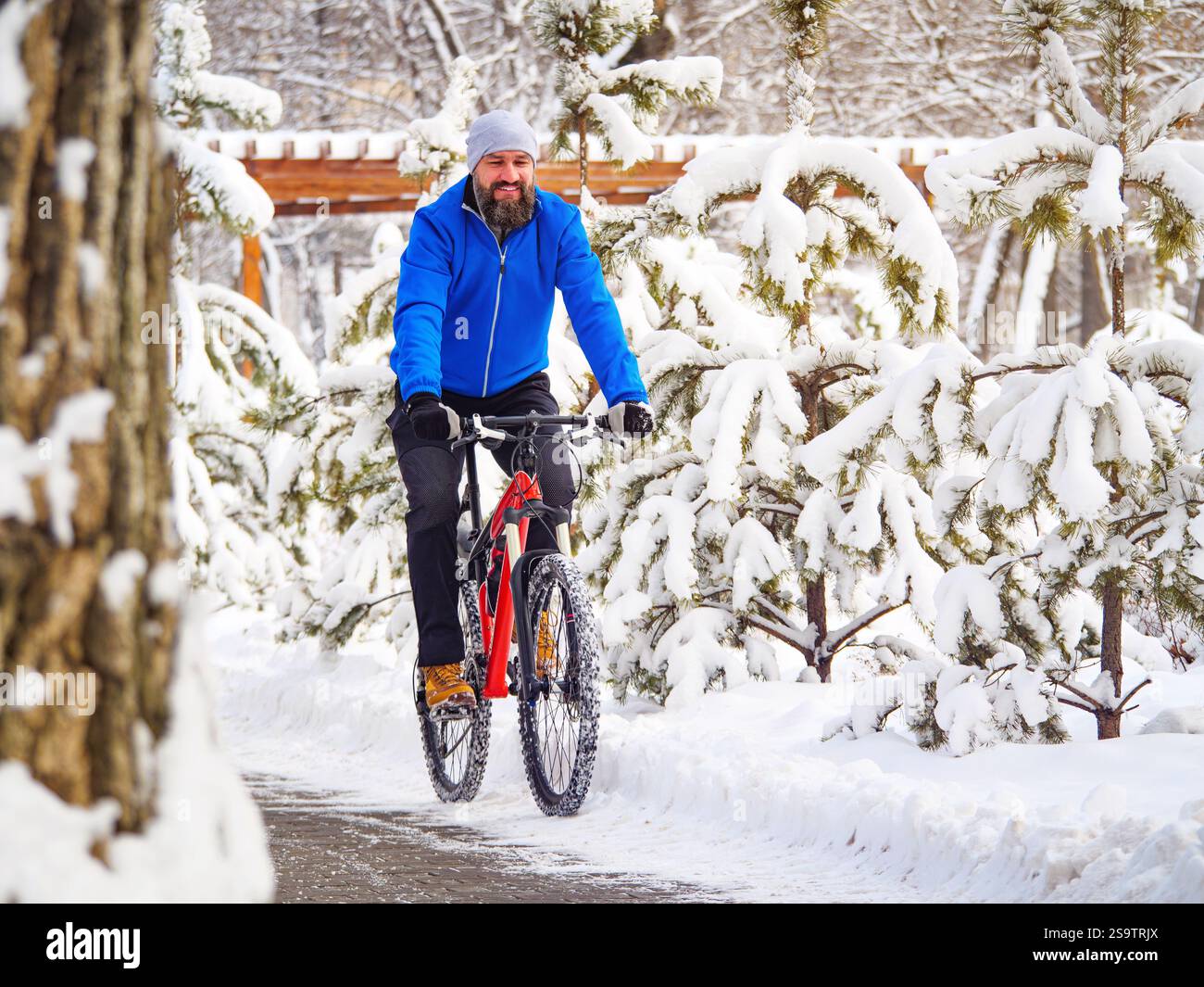 Un uomo gioioso cavalca una bicicletta in un parco invernale tra alberi innevati. Ciclista barbuto in una giacca blu.. Stile di vita attivo Foto Stock