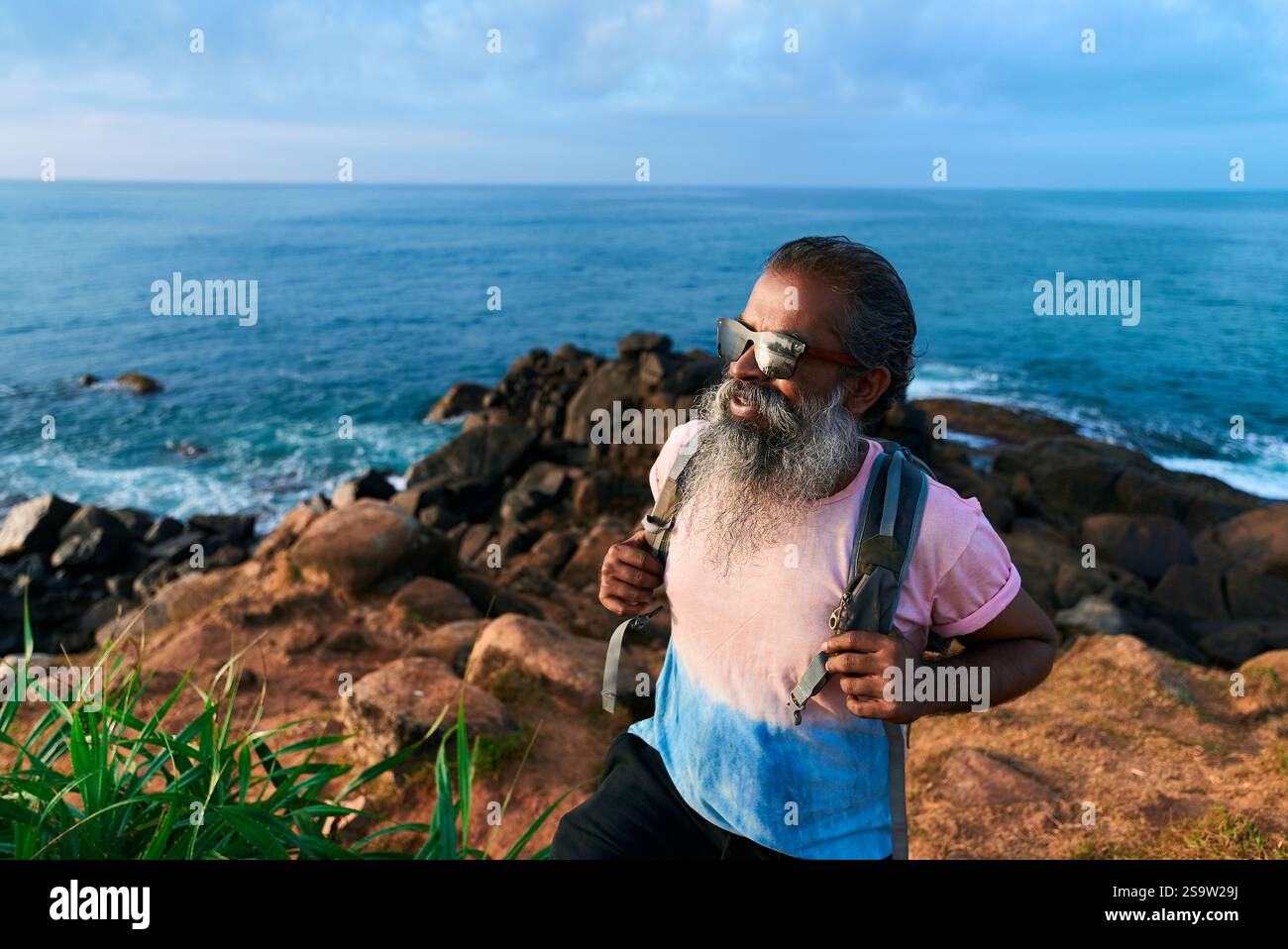 Elegante uomo anziano con barba, escursioni sul mare roccioso, occhiali da sole, zaino. Avventura attiva. Viaggio di pensionamento, destinazione costiera. Sano Foto Stock