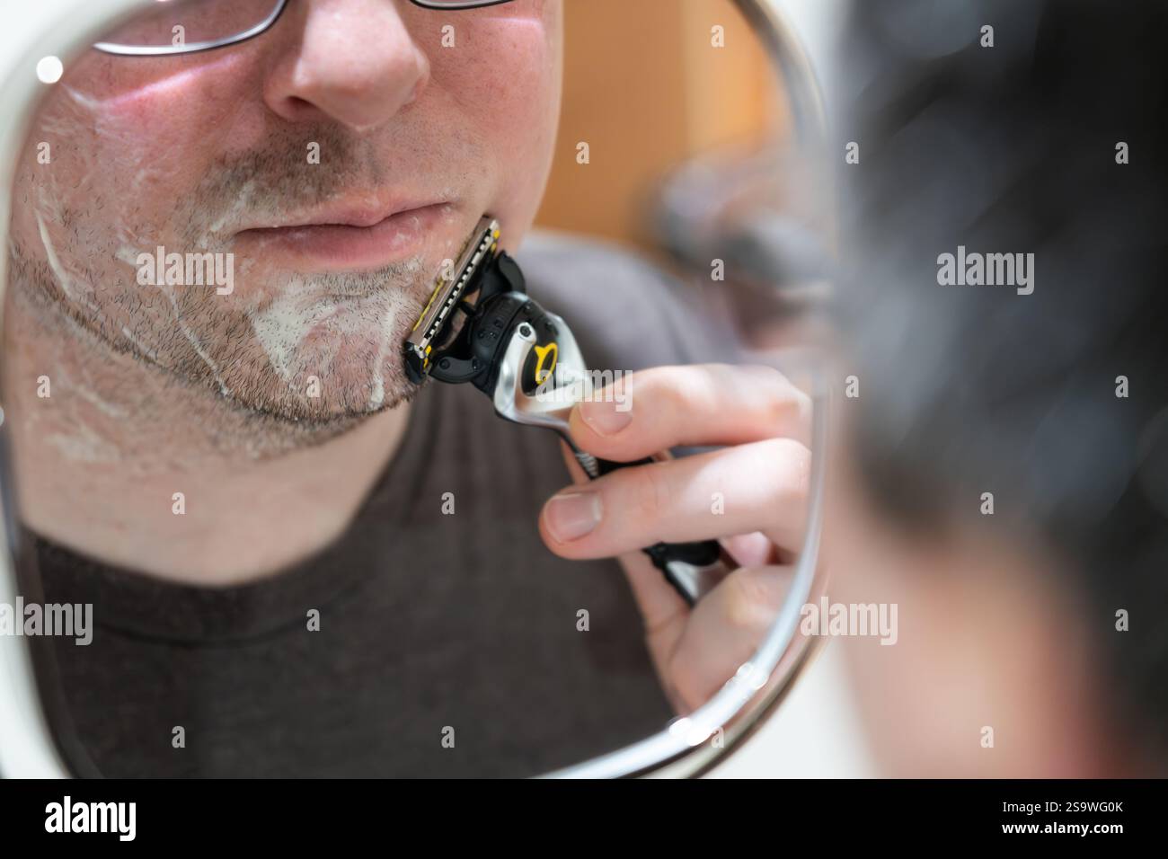 Uomo di mezza età che guarda in uno specchio allungabile montato a parete e si rade con un rasoio di sicurezza Gillette, Regno Unito. Concetto: Cura personale, routine mattutina Foto Stock