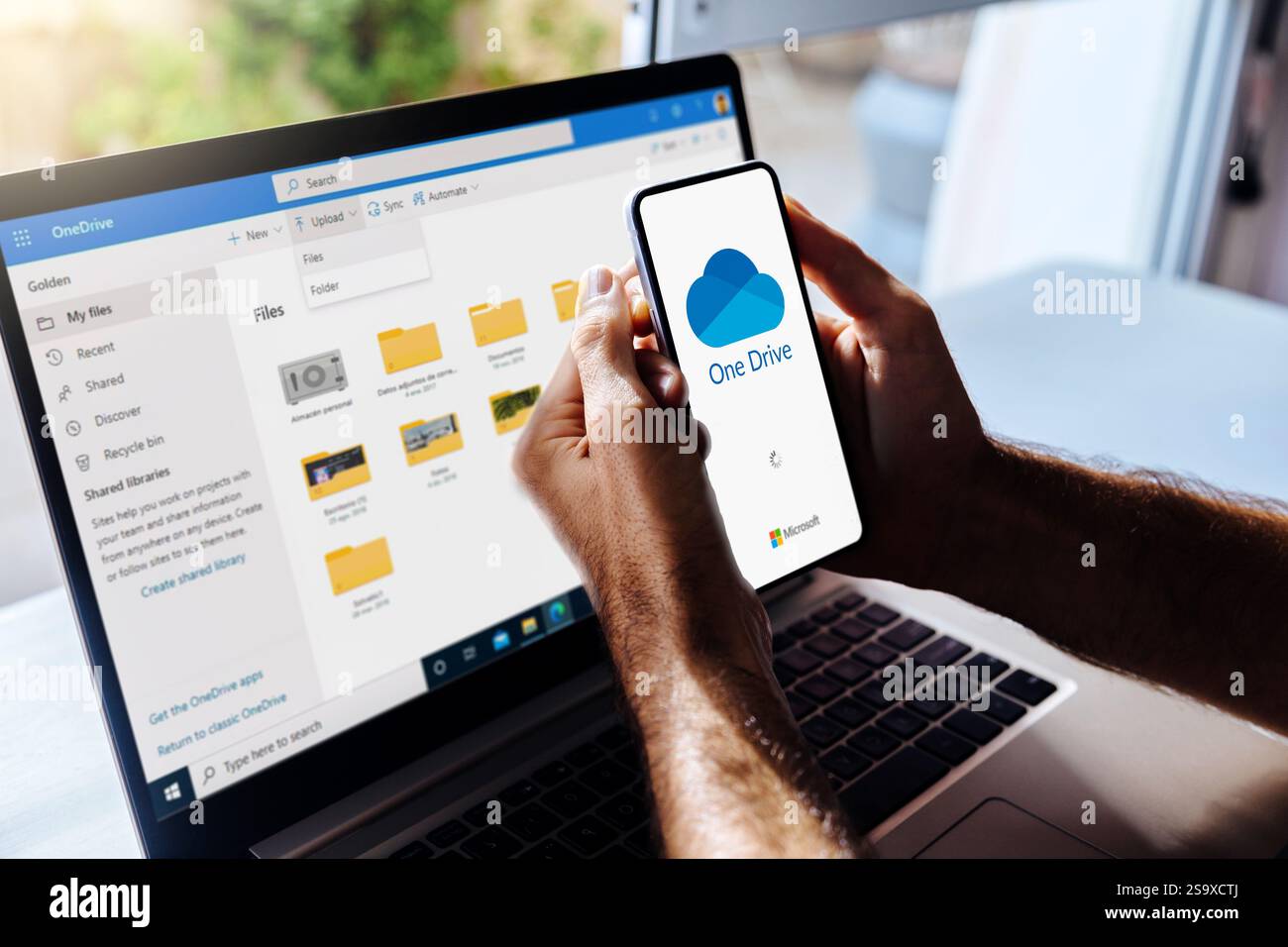 OneDrive sul telefono cellulare. Uomo con un telefono cellulare con l'app di archiviazione cloud Microsoft sullo schermo. Laptop sullo sfondo che mostra i file di un'unità Foto Stock