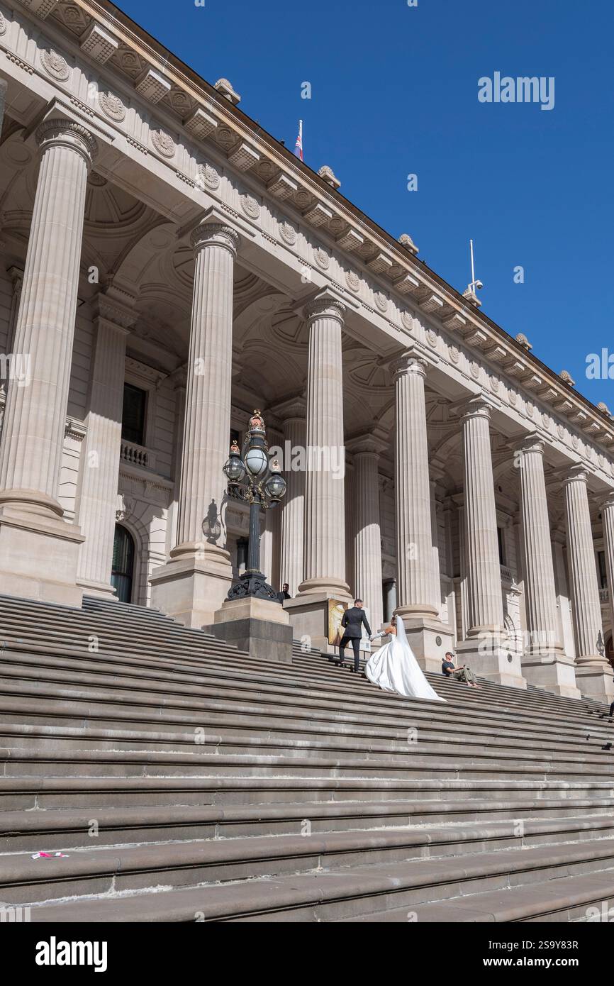Lo sposo in abito nero porta la sposa in un abito da sposa bianco sui gradini di pietra del Parlamento in una giornata di sole con il cielo blu a Melbourne in Australia Foto Stock
