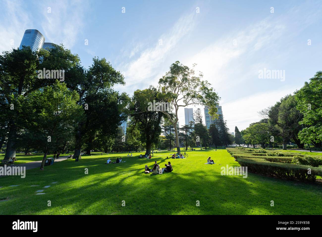Soleggiato nel tardo pomeriggio, le persone si siedono e si rilassano sotto il sole caldo sui prati dei Carlton Gardens, un parco nel nord di Melbourne dichiarato Patrimonio dell'Umanità Foto Stock