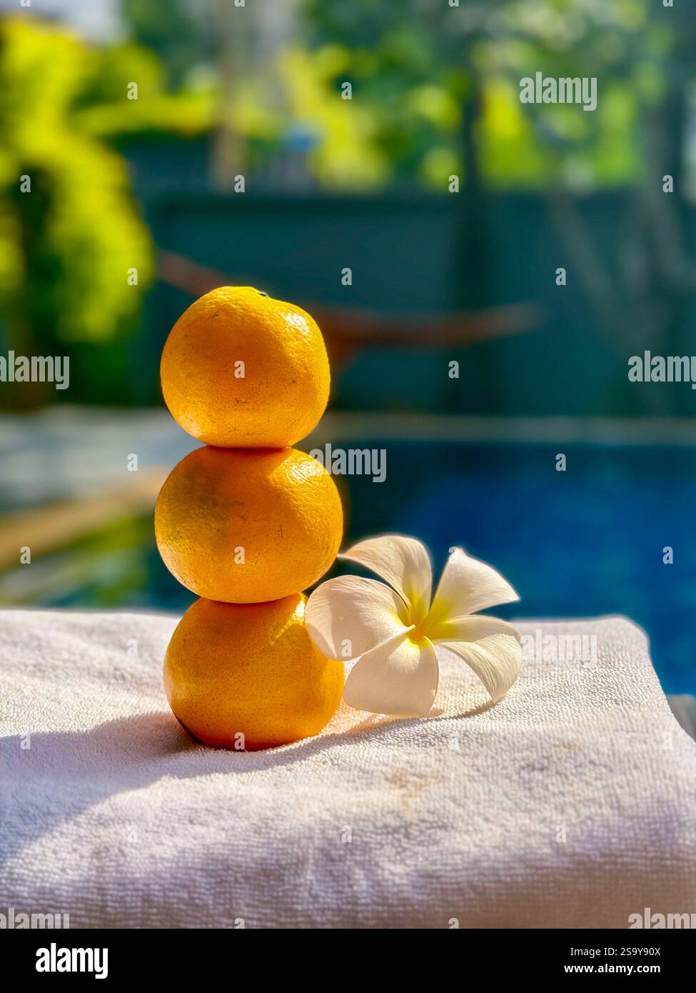 Arance sovrapposte con Fiore tropicale presso la piscina Una pila bilanciata di tre arance fresche accompagnate da un fiore tropicale bianco su un asciugamano, agains Foto Stock