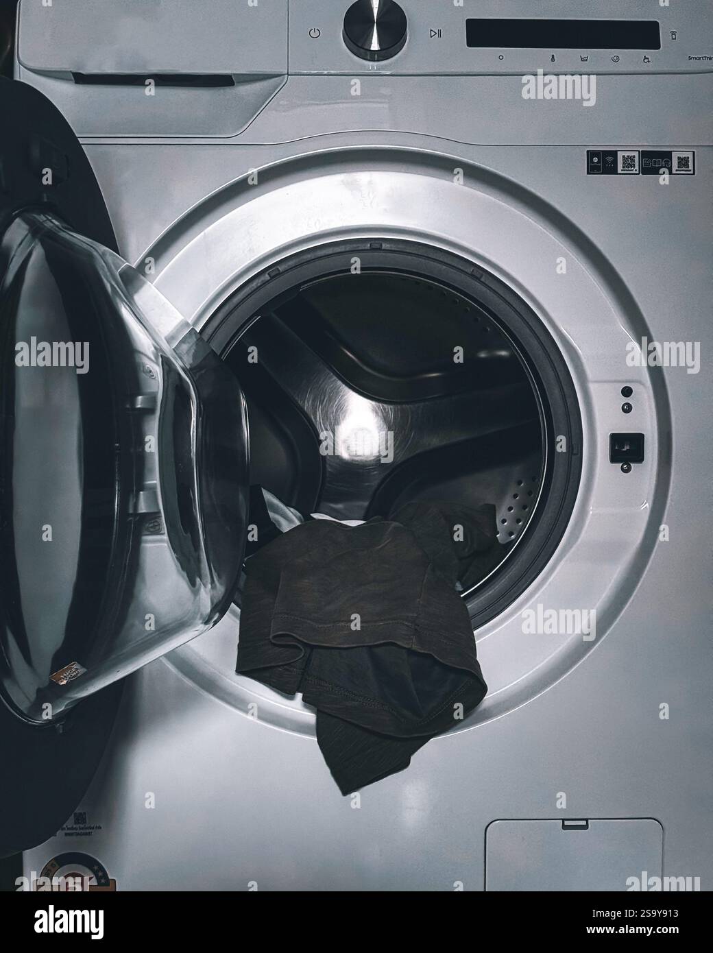 Lavatrice con sportello aperto Un primo piano di una lavatrice con lo sportello aperto, che mostra un indumento di colore scuro all'interno. Il tamburo metallico riflette t Foto Stock