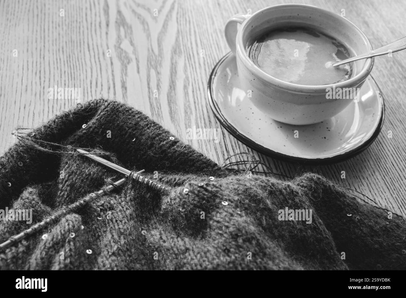 Maglione in maglia con paillettes e aghi per maglieria con tazza da caffè sul tavolo, bianco e nero. Sequenza in aghi con cappuccino, monocromatico. Foto Stock