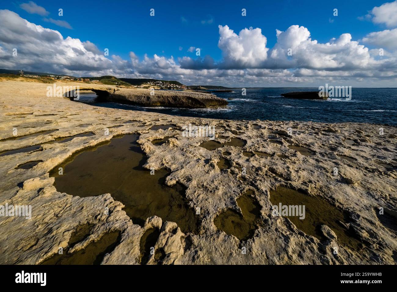Acqua piovana in piccoli fori d'acqua sulla costa rocciosa intorno all'Arco di S'Archittu dopo un tuono, cielo blu con nuvole bianche in lontananza. Foto Stock