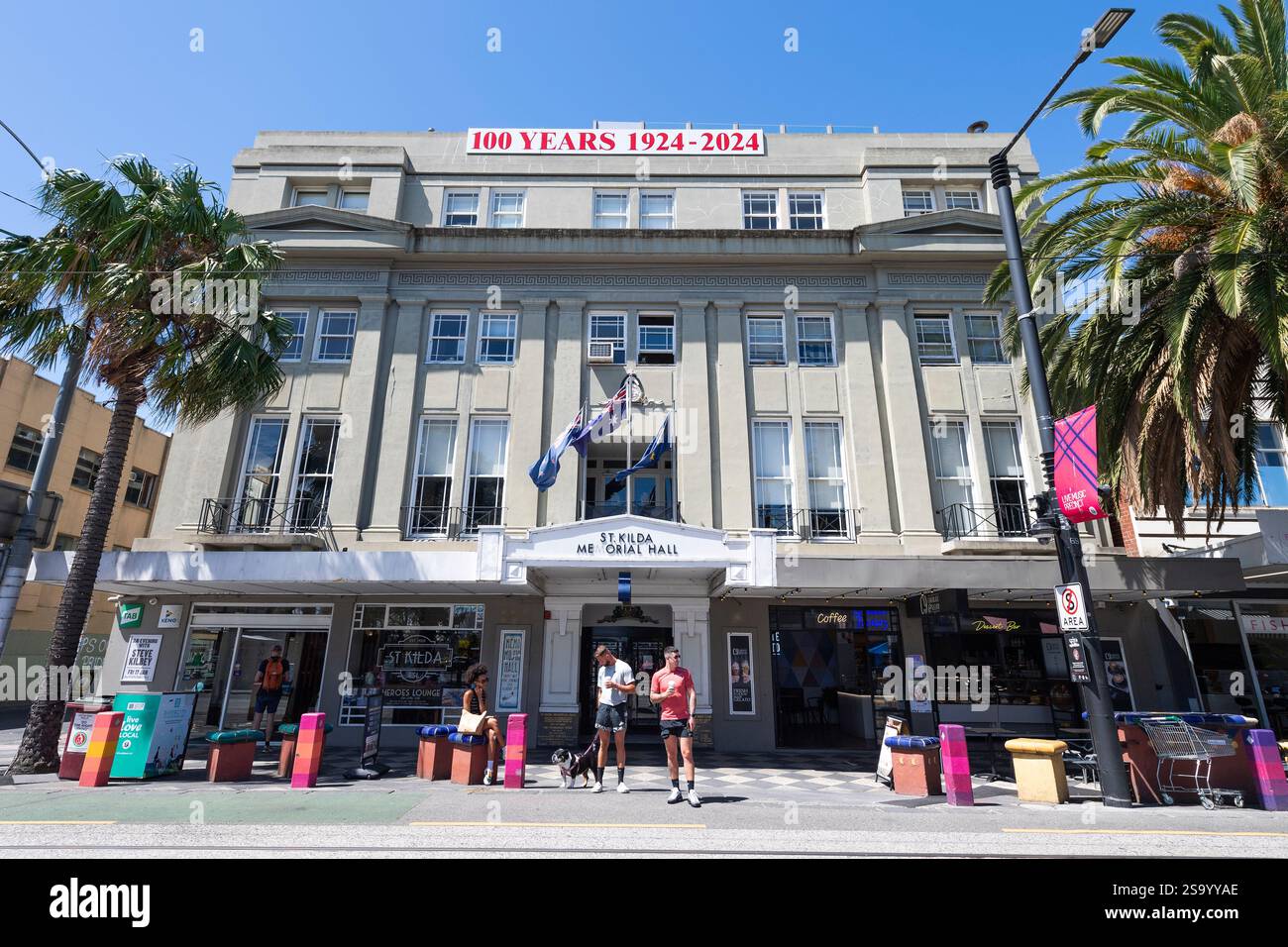 Due giovani uomini che indossano pantaloncini portano un cane davanti alla St Kilda Memorial Hall, Acland Street, Melbourne, Australia, in una soleggiata giornata estiva con il cielo blu. Foto Stock