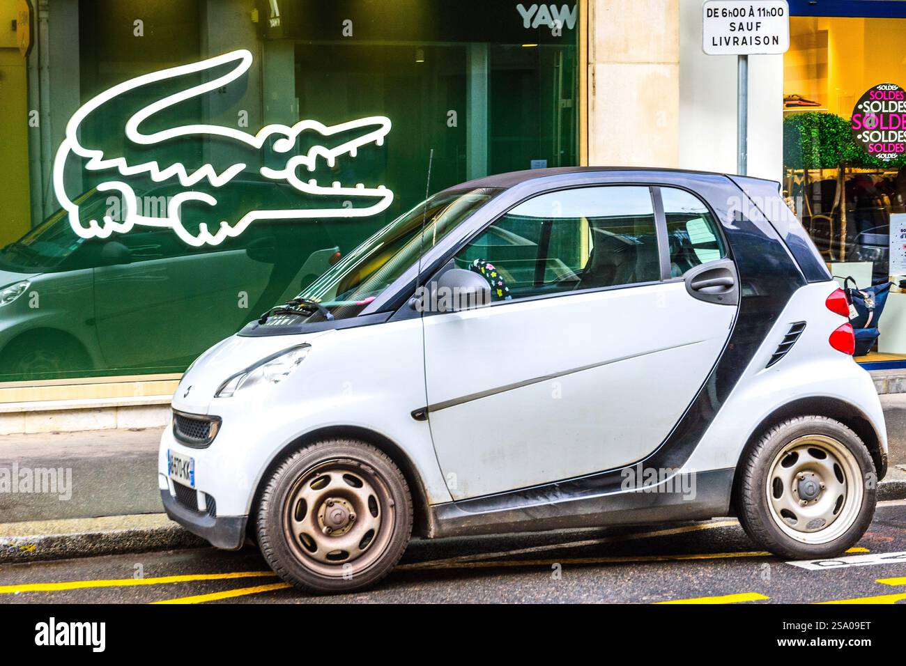 Un minicar 'Smart ForTwo' parcheggiato in una strada cittadina di fronte alla vetrina del negozio con il simbolo 'Lacoste' visualizzato - Tours, Indre-et-Loire (37), Francia. Foto Stock