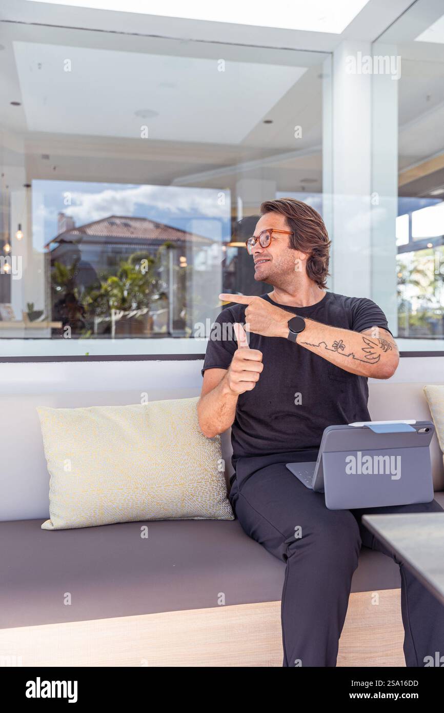Un uomo hipster felice regala i pollici verso l'alto, punta a sinistra mentre lavora in ufficio, foto verticale. Foto Stock