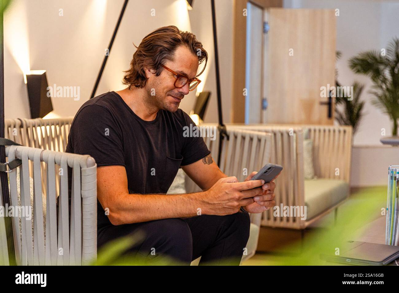 Un uomo è seduto su un divano e guarda il suo cellulare. La camera è ben illuminata e presenta un design moderno Foto Stock