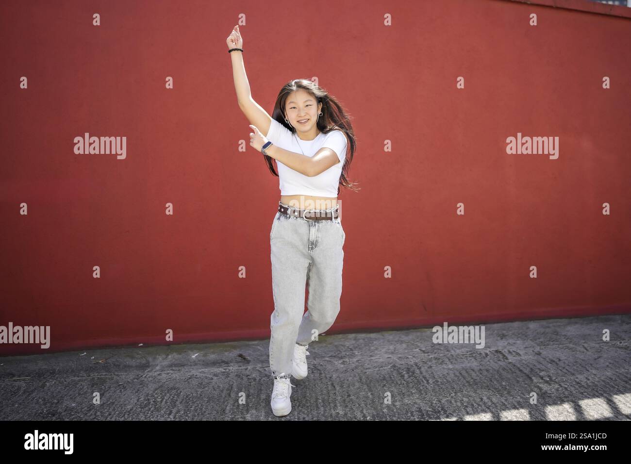 Energica donna asiatica che salta danzando la danza urbana K-pop in città contro un muro rosso urbano Foto Stock