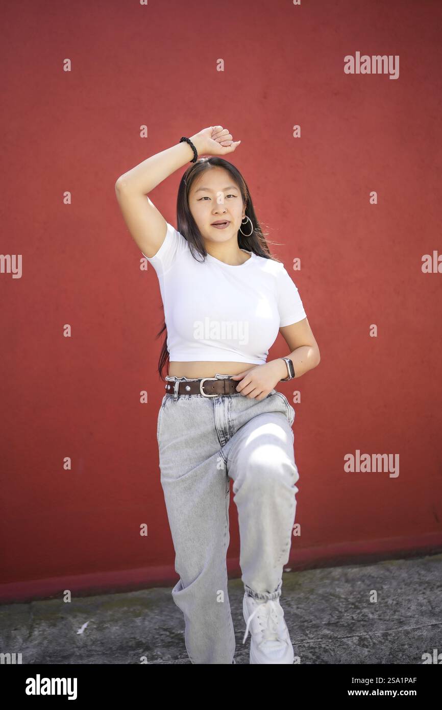 Foto verticale di una bella adolescente asiatica che danza k-pop per strada contro un muro rosso Foto Stock