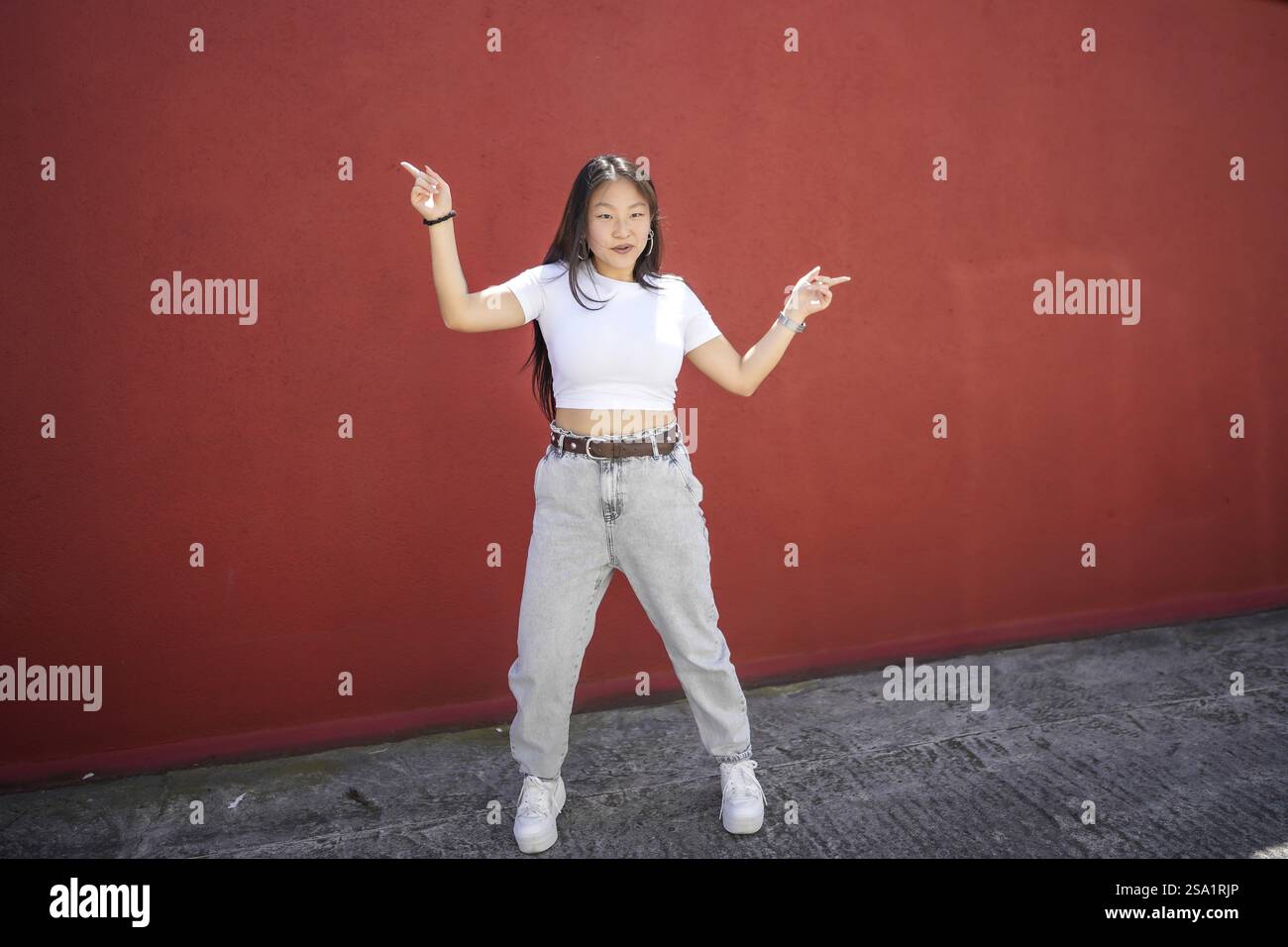 Foto a figura intera di una giovane ballerina asiatica energica che esegue danza urbana K-pop in città contro un muro rosso Foto Stock