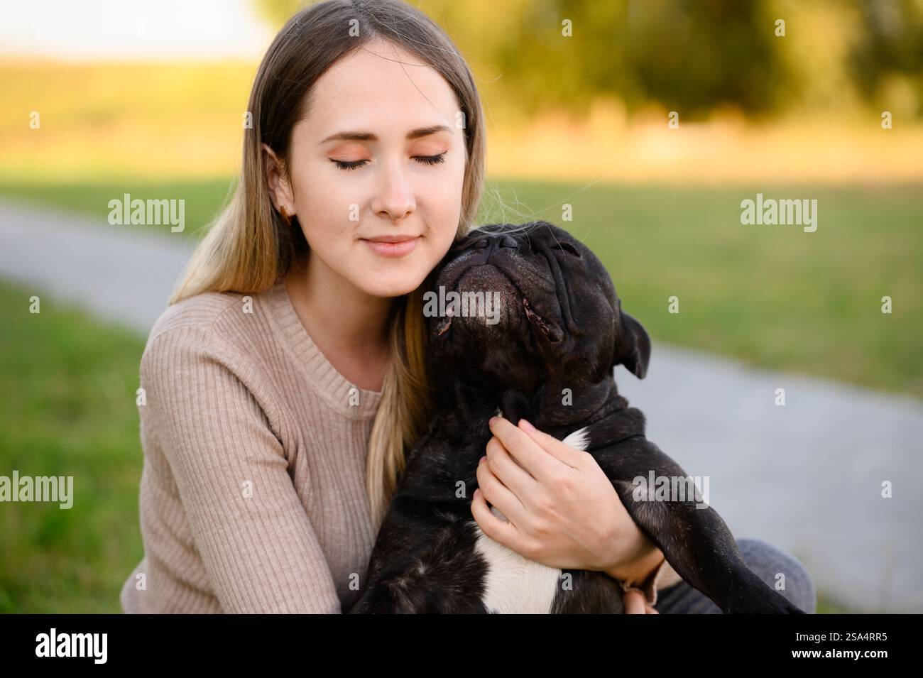 La giovane donna abbraccia, coccola il suo amato bulldog francese, sorride, chiude gli occhi mentre cammina. Il cane preferito tra le braccia del proprietario. Concetto di cura, adozione, cura e amore per gli animali domestici. Foto Stock