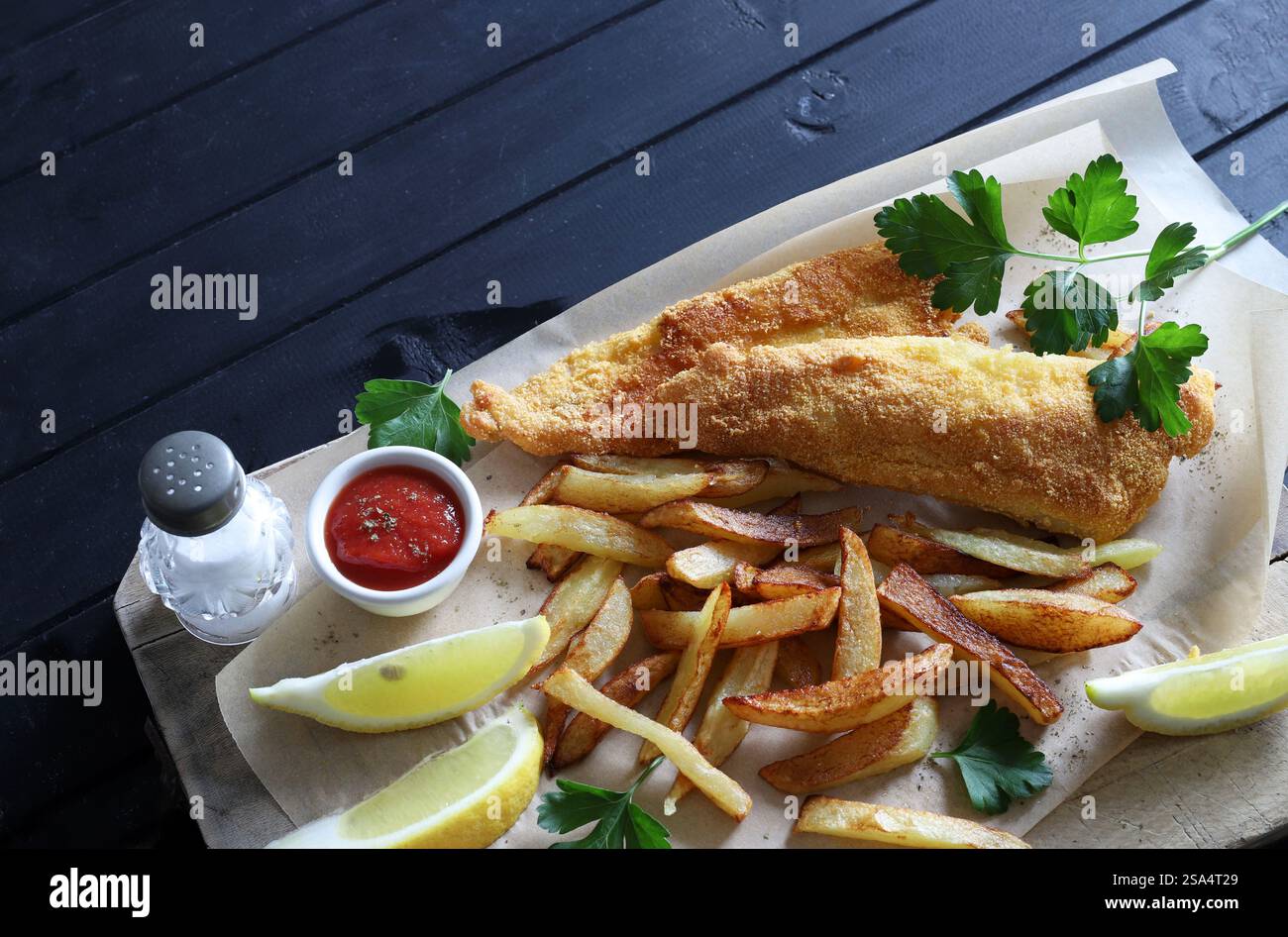 Pesce fritto e patatine fritte servite con salsa e limone a fette nel piatto su sfondo nero di legno. Piatto di pesce britannico. Direttamente sopra. Foto Stock