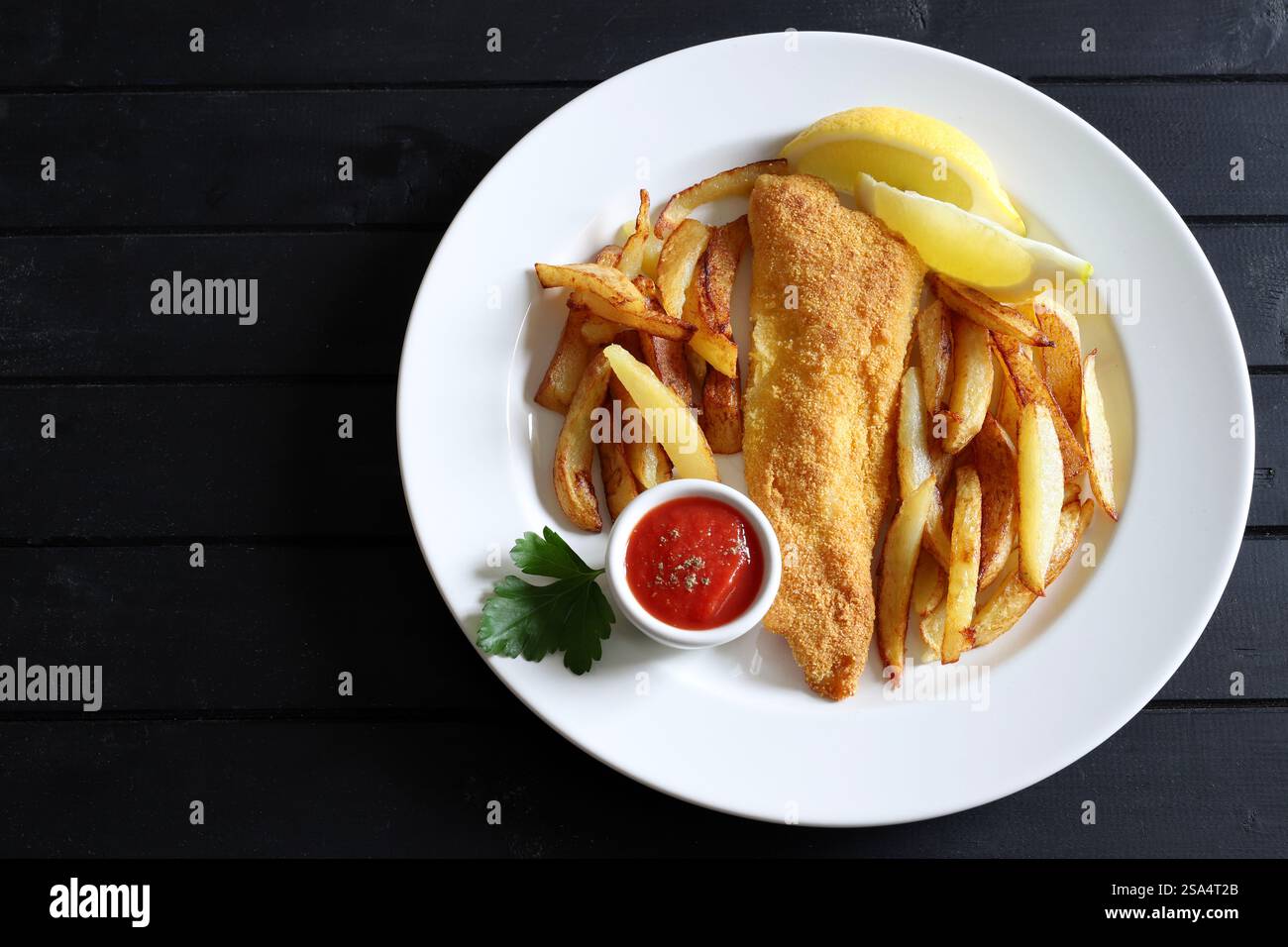 Pesce fritto e patatine fritte servite con salsa e limone a fette nel piatto su sfondo nero di legno. Piatto di pesce britannico. Direttamente sopra. Foto Stock
