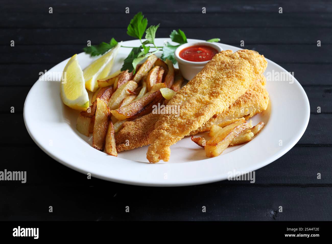 Pesce fritto e patatine fritte servite con salsa e limone a fette nel piatto su sfondo nero di legno. Piatto di pesce britannico. Direttamente sopra. Foto Stock