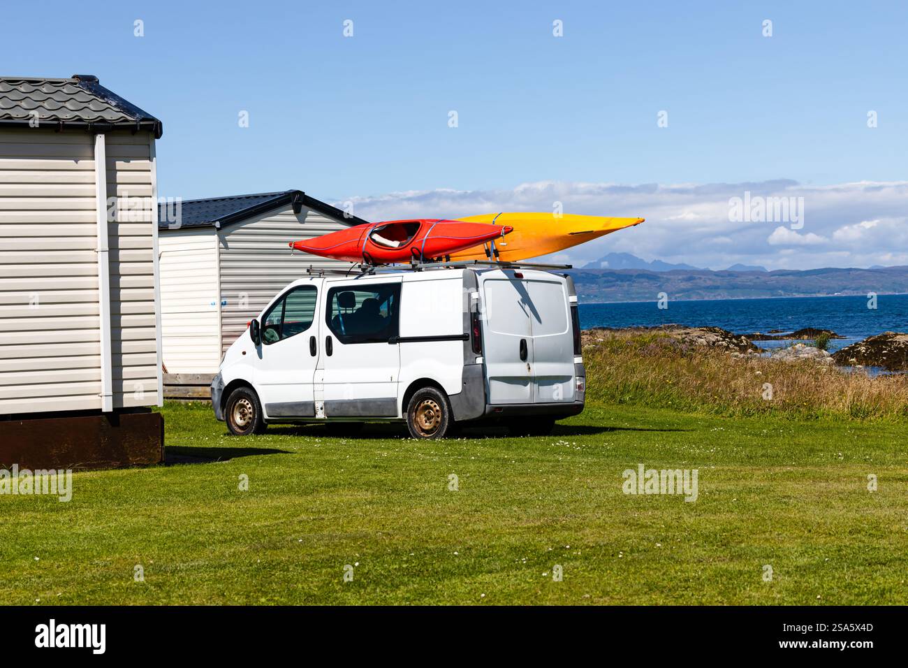 un vecchio furgone bianco parcheggiato sull'erba con 2 canoe legate al portapacchi in una giornata di sole. Mare colline, montagne sullo sfondo Foto Stock