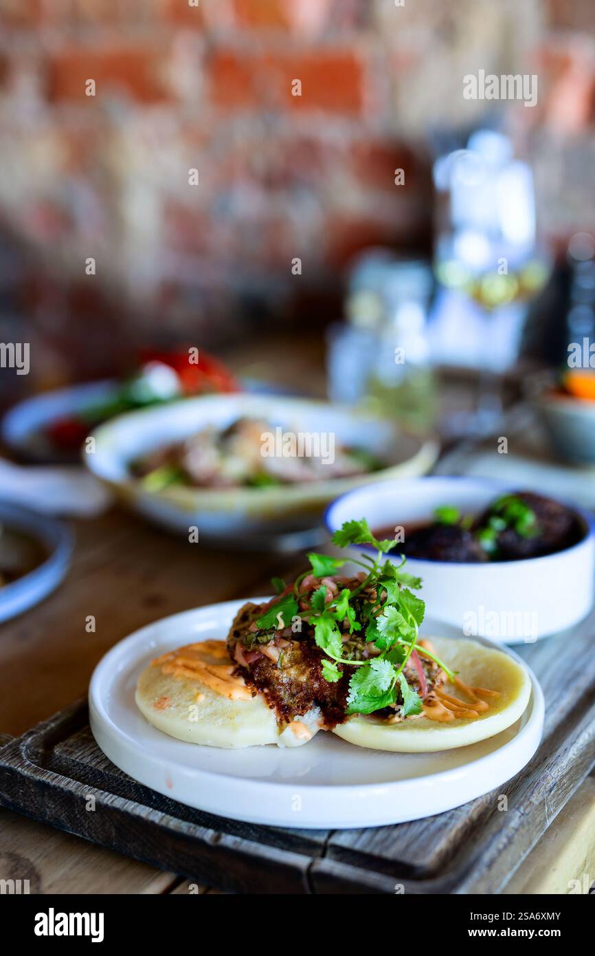 Un delizioso piatto bao Bun caratterizzato da ripieno croccante, coriandolo fresco e una salsa cremosa, servito su un piatto bianco con uno sfondo rustico da tavolo in legno. Foto Stock