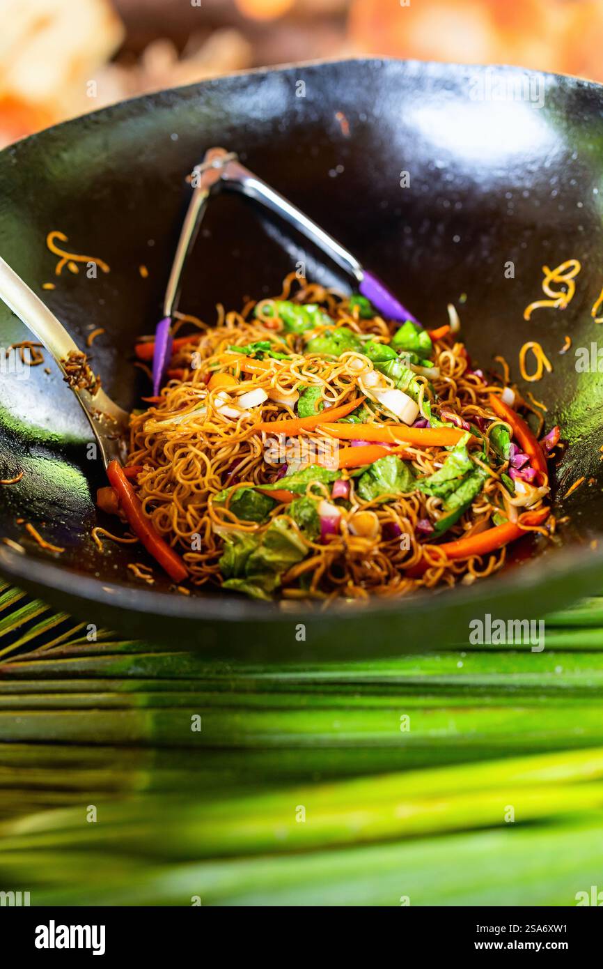Un piatto vibrante di spaghetti saltati con verdure fresche come carote, verdure a foglia verde e cipolle, servito in un wok nero con pinze e un cucchiaio da portata Foto Stock