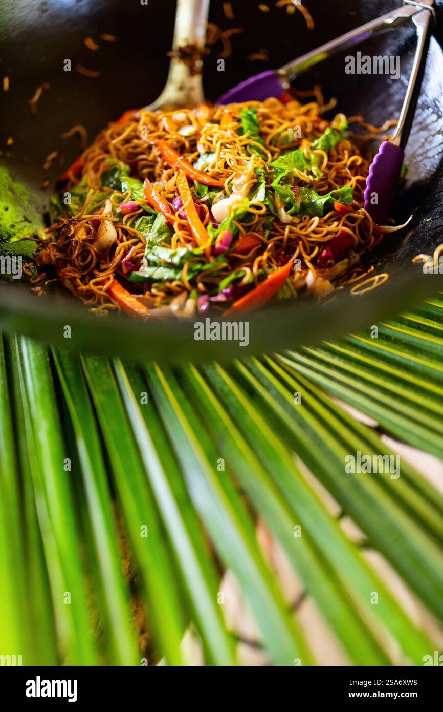 Un piatto vibrante di spaghetti saltati con verdure fresche come carote, verdure a foglia verde e cipolle, servito in un wok nero con pinze e un cucchiaio da portata Foto Stock