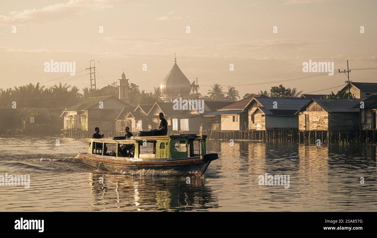Una nave passeggeri Kelotok che trasporta i turisti lungo il fiume Martapura con un villaggio tradizionale e una moschea sullo sfondo Foto Stock