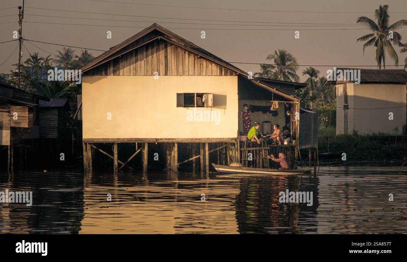 una palafitta accanto ad un fiume dove due donne interagiscono con un'altra donna su una barca. Foto Stock
