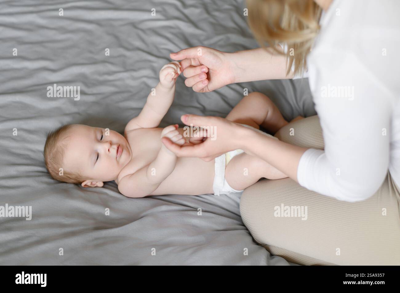 La mamma si siede sul letto e gioca con il bambino sdraiato sul letto, tenendosi tenero le mani. Alle donne piace la maternità. Concetto di famiglia, amore, cura di un bambino, Foto Stock