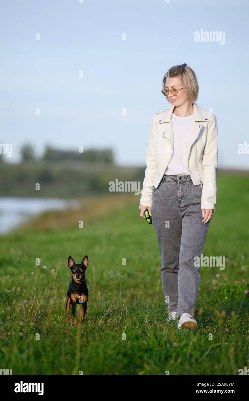 Una donna con gli occhiali cammina con un giocattolo terrier russo dai capelli lisci al guinzaglio in un parco vicino a uno stagno in una soleggiata giornata estiva, divertendosi all'aria aperta. Frie Foto Stock