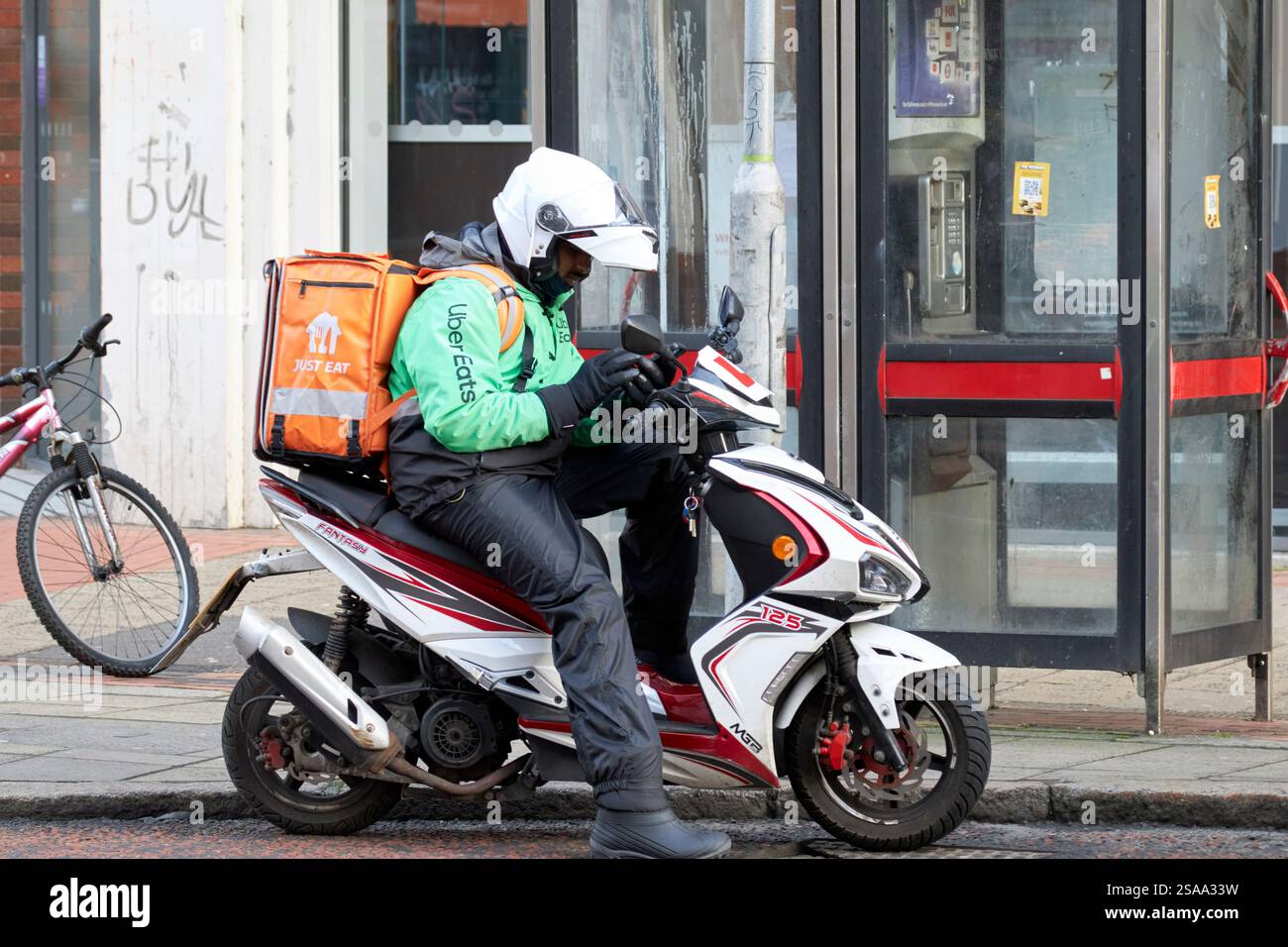 mangia solo un addetto alla consegna seduto su un ciclomotore nel centro di belfast guardando il suo telefono belfast, irlanda del nord, regno unito Foto Stock