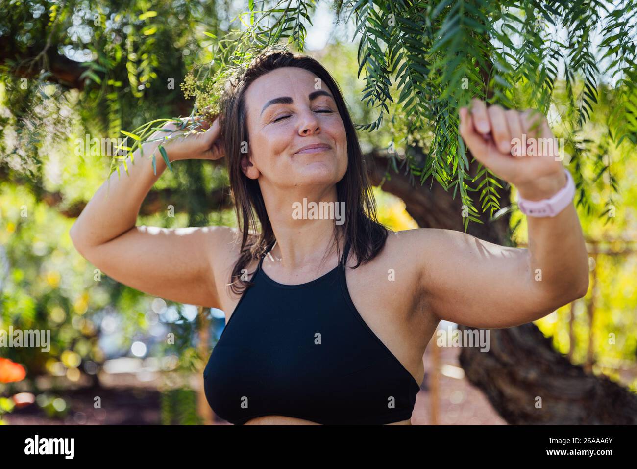 Una donna spensierata è in piedi in un parco, sorride e respira l'aria fresca, amante della natura. Sta indossando una canotta nera e si sta godendo l'uscita Foto Stock