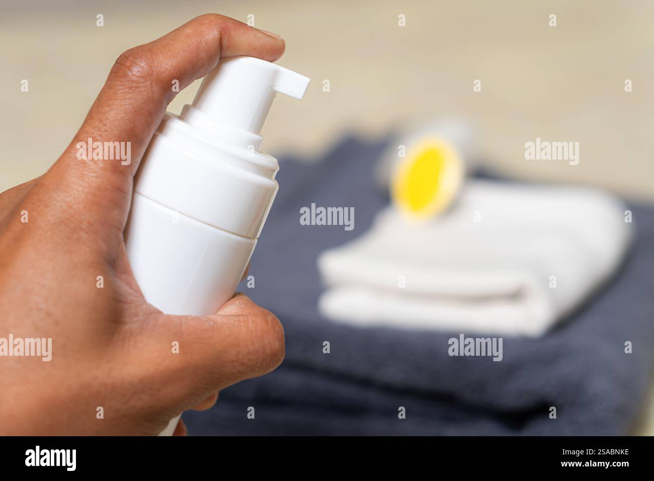 Una mano che regge un flacone bianco, pronto per erogare un prodotto. Sullo sfondo sono presenti asciugamani ben piegati, uno bianco e uno grigio scuro e un Foto Stock