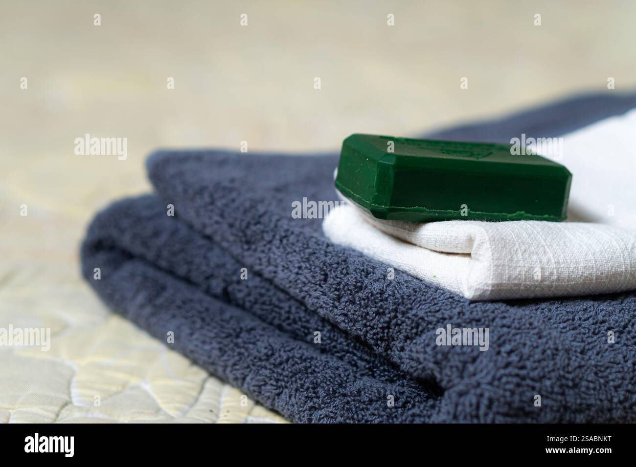Una pila di asciugamani ben piegati nei colori grigio scuro e bianco, con una barra verde di sapone posta sopra. Lo sfondo presenta un morbido trapuntato sfocato Foto Stock