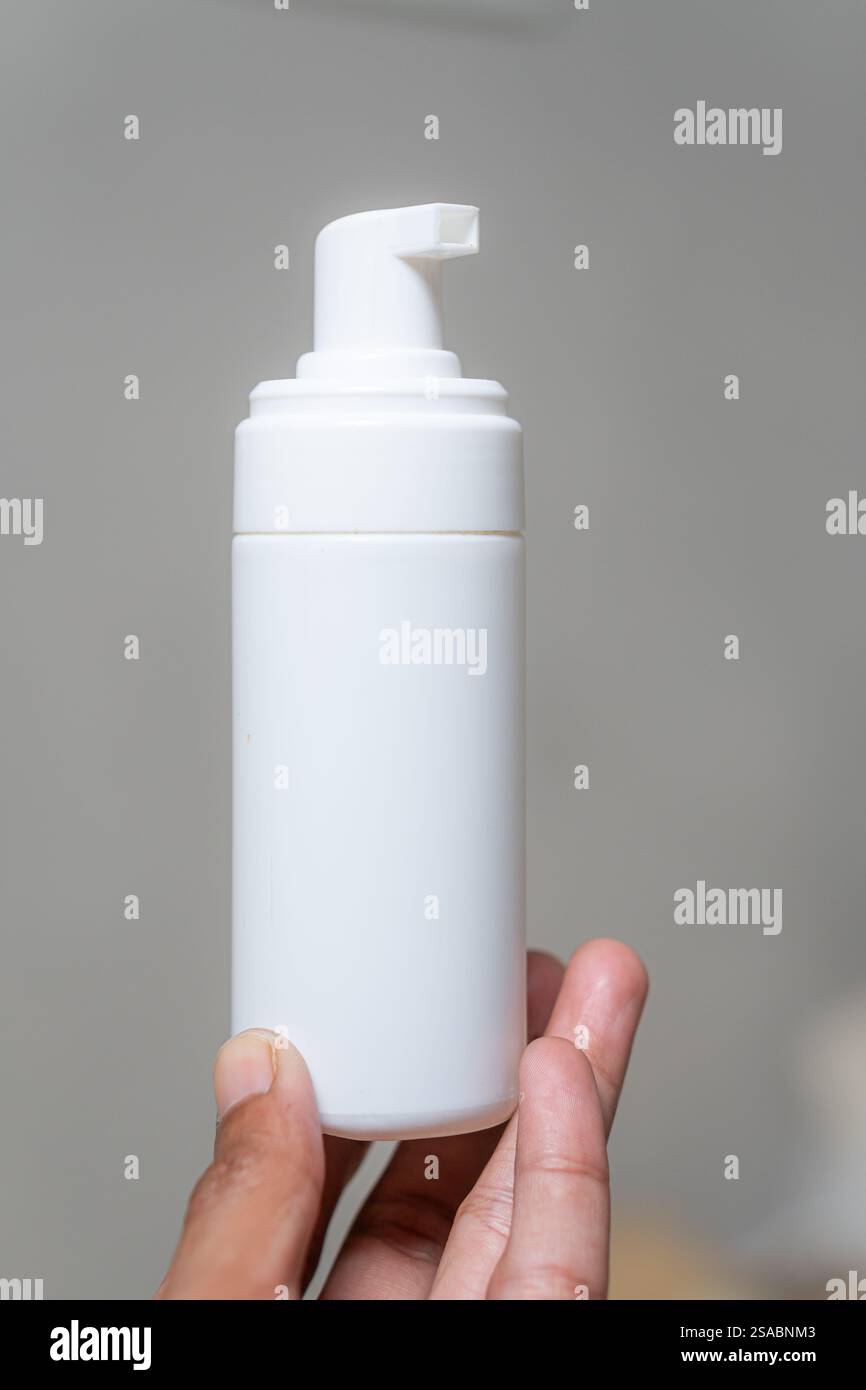Una persona che regge un flacone bianco su uno sfondo neutro. Il flacone è cilindrico con un dispenser a pompa sulla parte superiore, adatto per lozioni o cre Foto Stock