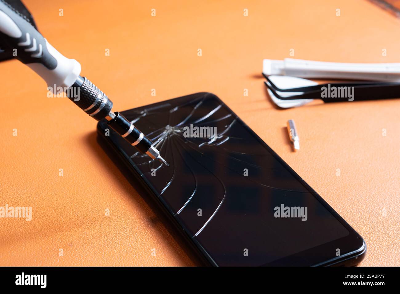 Primo piano di un set di cacciaviti di precisione con punte multiple, un portautensili giallo e uno smartphone con schermo incrinato su una superficie arancione. Foto Stock