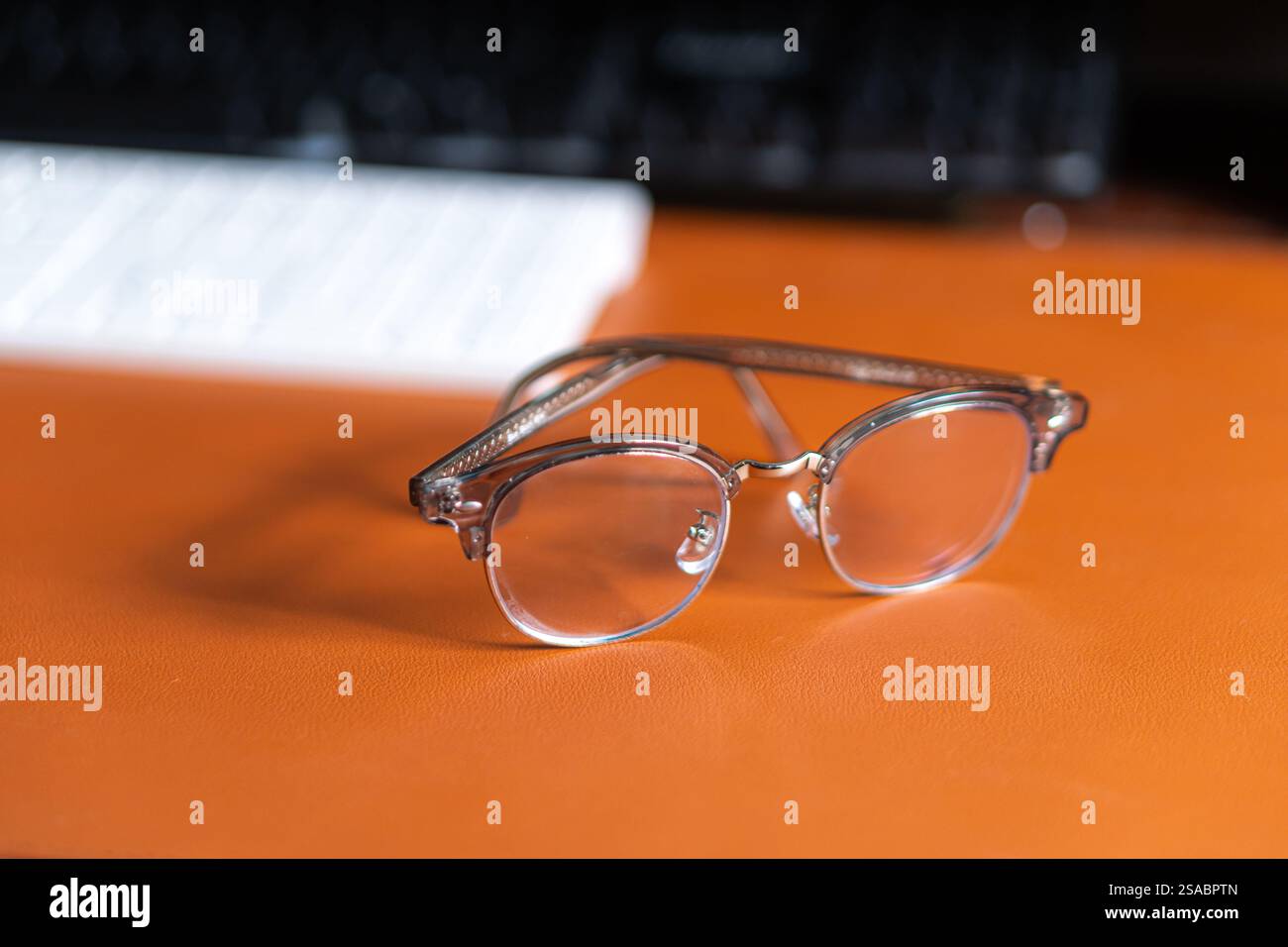 Un paio di eleganti occhiali da vista appoggiati su una superficie in pelle marrone, con una tastiera sfocata sullo sfondo. Foto Stock