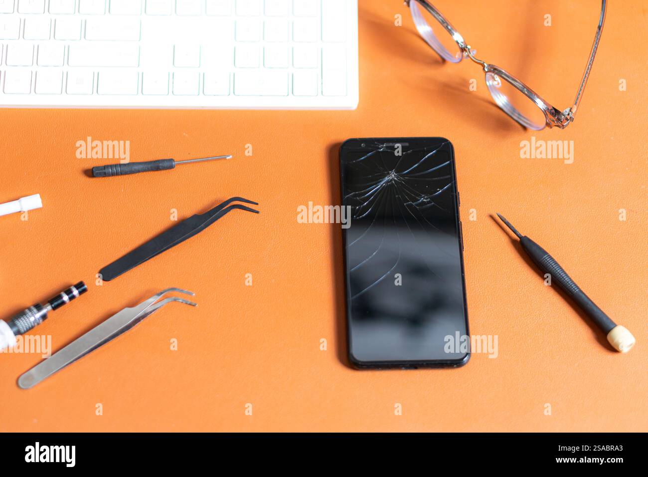 Uno smartphone con uno schermo incrinato posizionato su una scrivania arancione. Gli strumenti circostanti includono pinzette, un cacciavite e una tastiera, insieme a un paio di Foto Stock