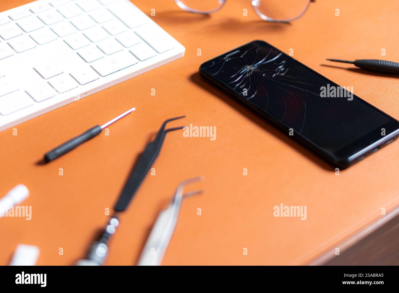 Uno smartphone con uno schermo incrinato posizionato su una scrivania arancione. Gli strumenti circostanti includono pinzette, un cacciavite e una tastiera, insieme a un paio di Foto Stock