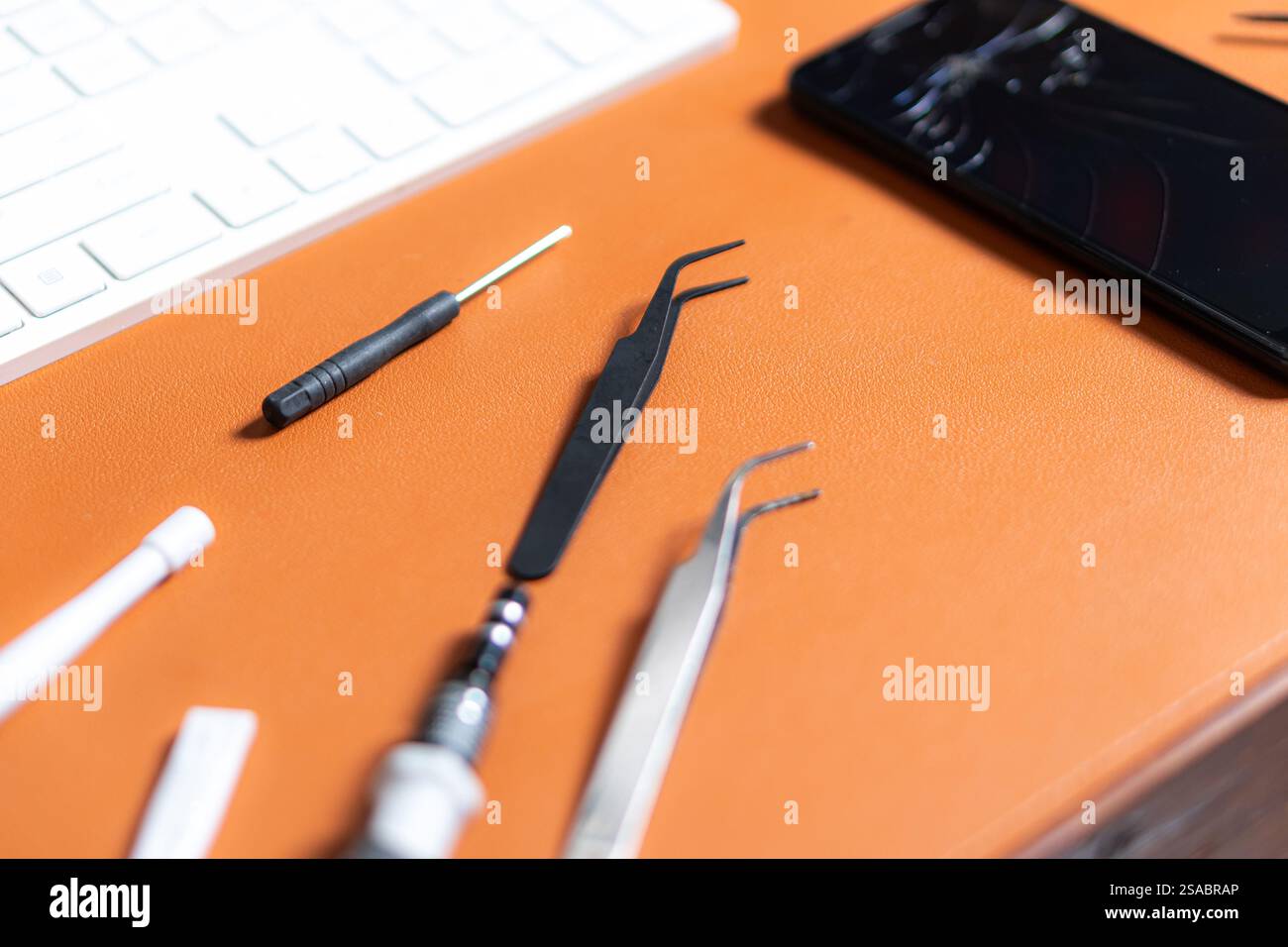 Uno smartphone con uno schermo incrinato posizionato su una scrivania arancione. Gli strumenti circostanti includono pinzette, un cacciavite e una tastiera, insieme a un paio di Foto Stock