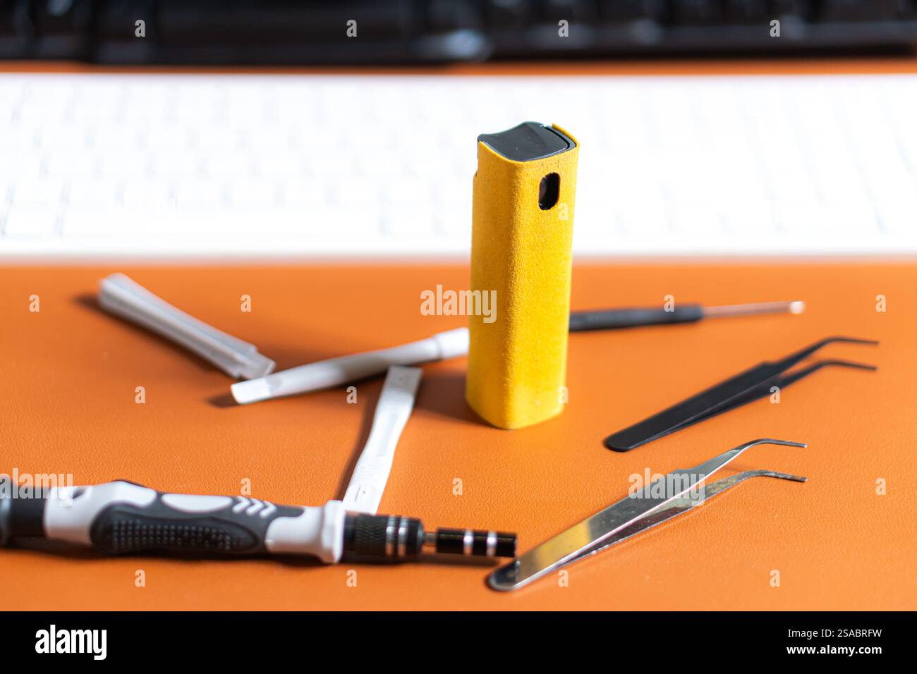 Una raccolta di strumenti tra cui un dispositivo giallo, cacciaviti e strumenti di precisione su una superficie arancione, con una tastiera sullo sfondo. Foto Stock