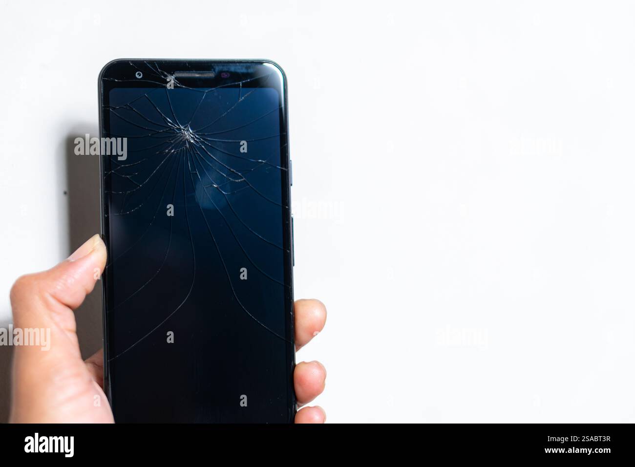 Primo piano di una mano che tiene in mano uno smartphone con uno schermo incrinato su uno sfondo bianco. Lo schermo è scuro e mette in evidenza il tipo ragnatela Foto Stock