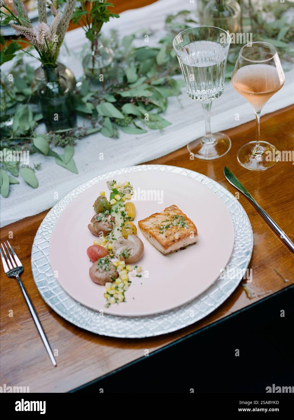 Un raffinato ambiente da tavola mette in mostra un piatto di carni e verdure ben organizzato insieme a un bicchiere di vino rosato. Il fogliame decorativo migliora l'AT Foto Stock