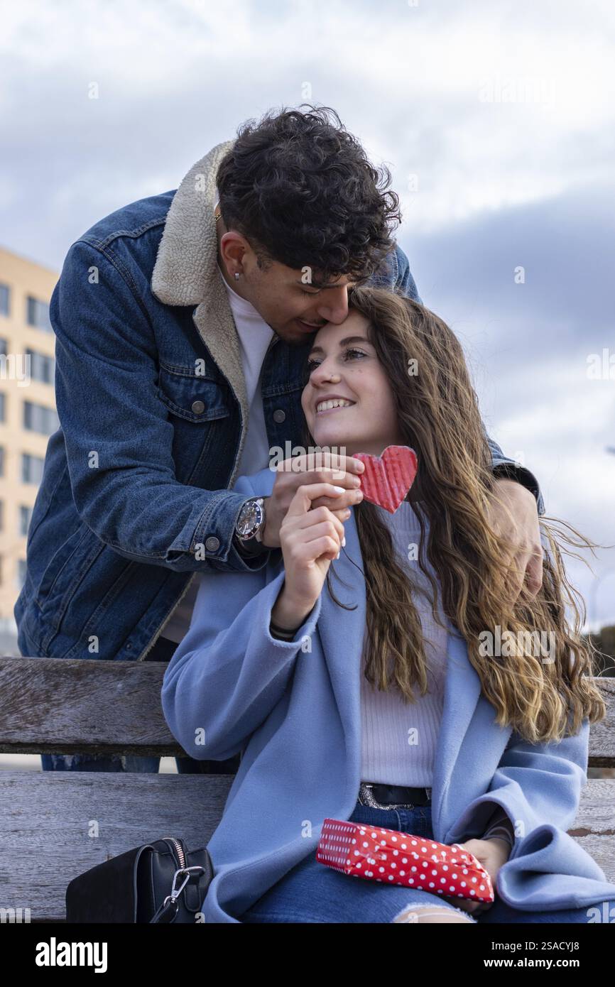 Ritratto verticale dell'uomo latino che abbraccia e bacia la sua ragazza da dietro, dandole una sorpresa con un dono e un cuore. celebrazione di uno speciale Foto Stock