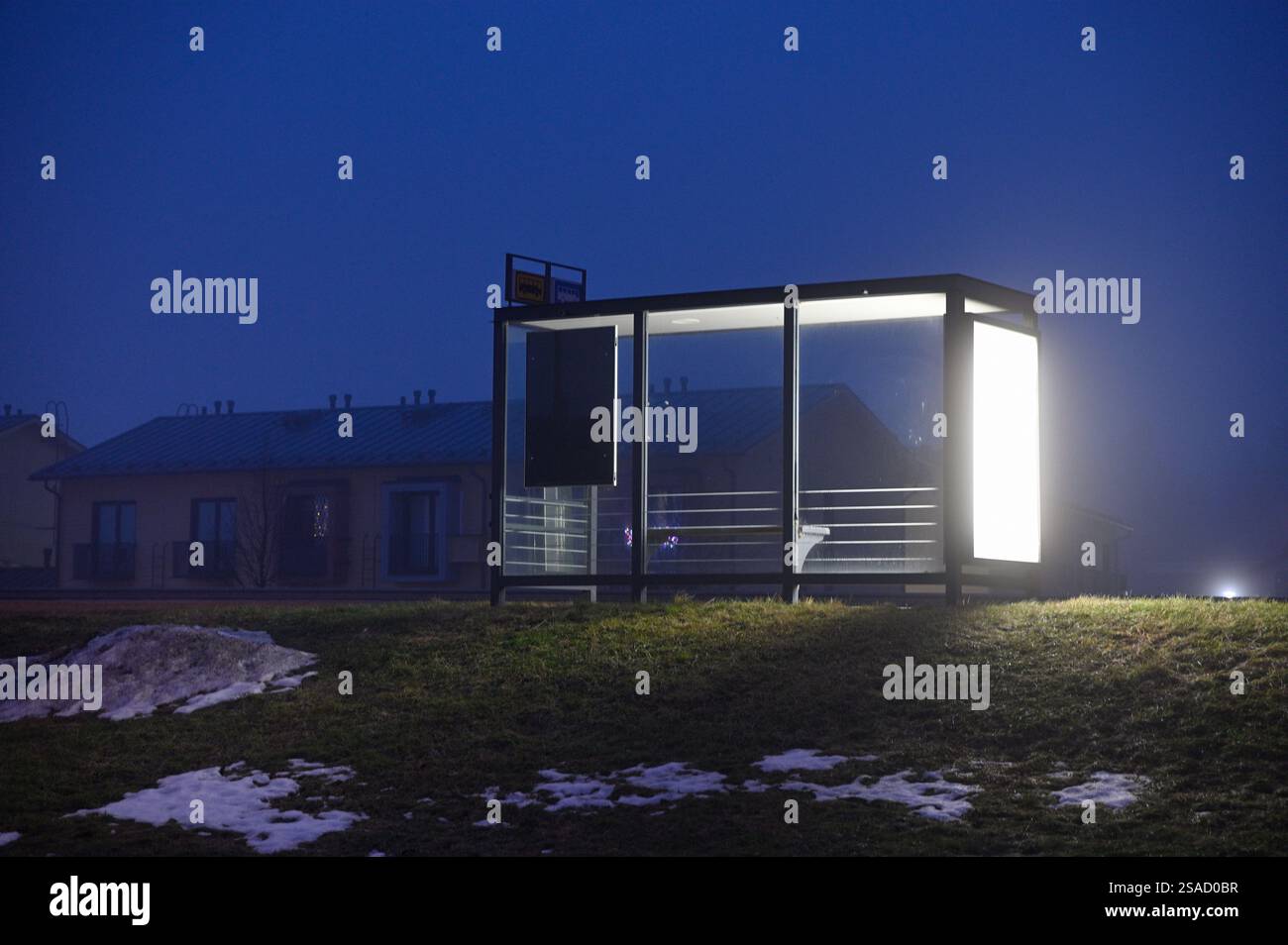 Una fermata minimalista dell'autobus illuminata da pannelli luminosi in una serata nebbiosa, immersa in un tranquillo paesaggio suburbano. Macchie di neve a terra e luce soffusa Foto Stock