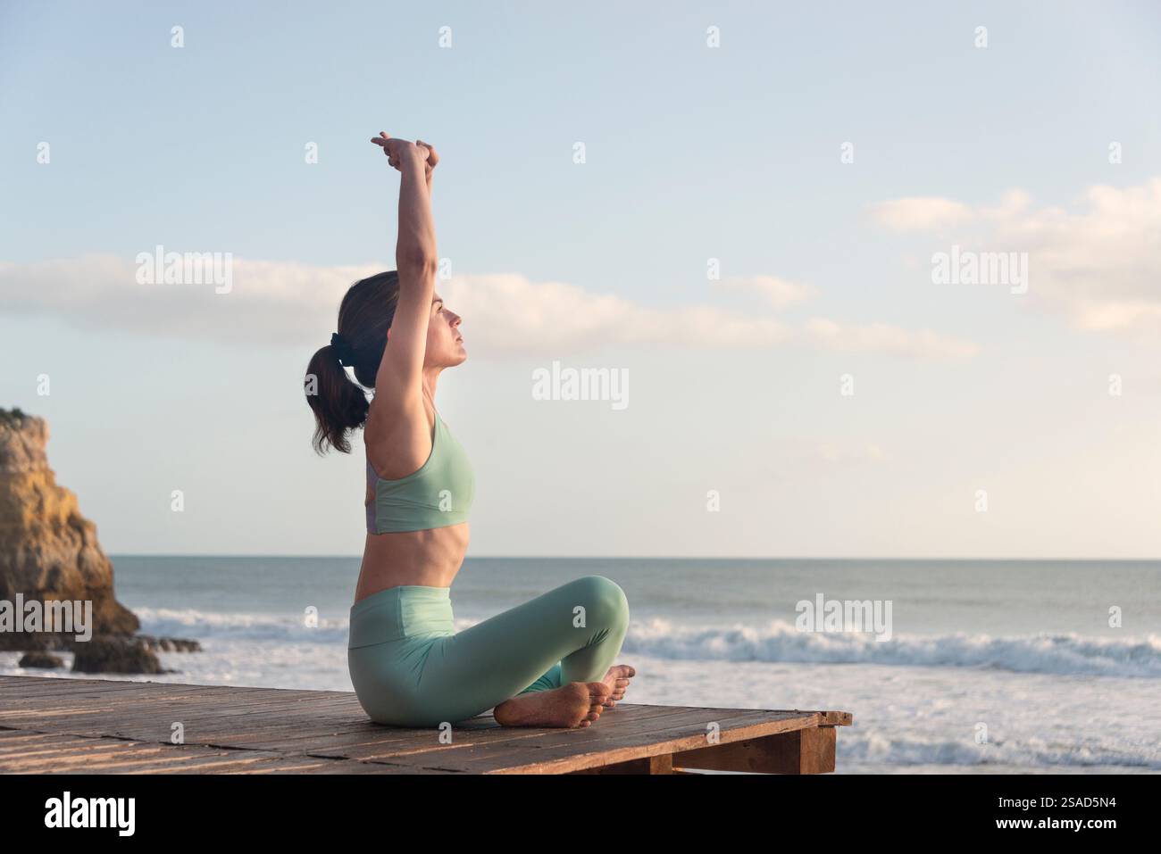 indossa una donna sportiva seduta accanto all'oceano che si allunga con le braccia sopra la testa Foto Stock