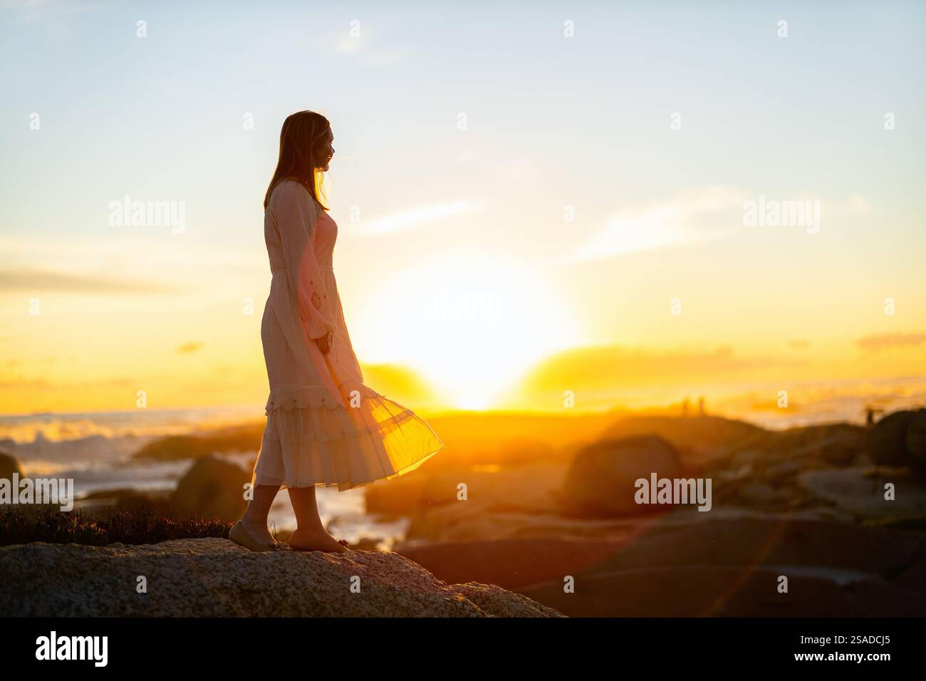 Una donna che indossa un abito bianco che scorre si erge su una costa rocciosa al tramonto, crogiolandosi di luce dorata mentre le onde dolci e un bagliore morbido creano una costa serena Foto Stock