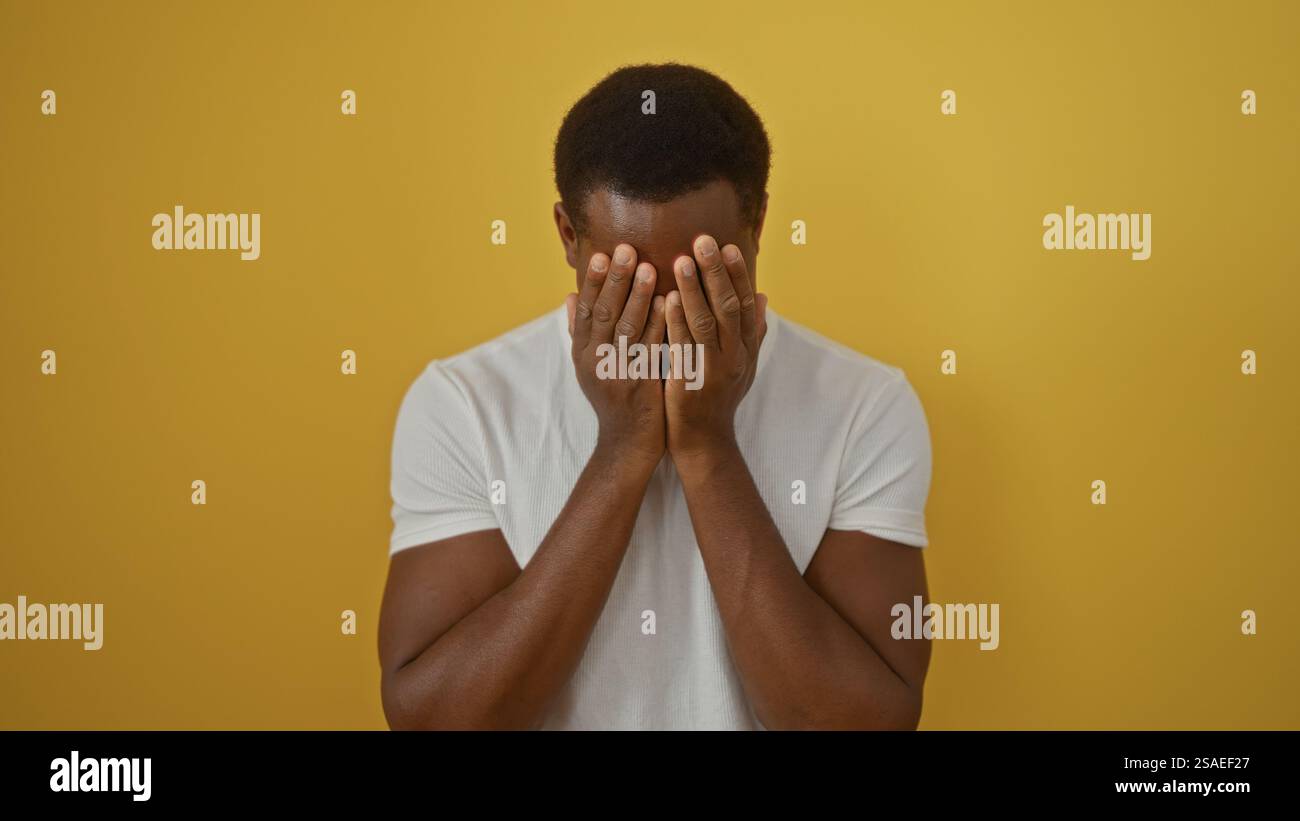 Il giovane uomo che ricopre il viso su sfondo giallo mostra emozione e contemplazione, enfatizzando la solitudine e l'introspezione in un ambiente a contrasto luminoso Foto Stock