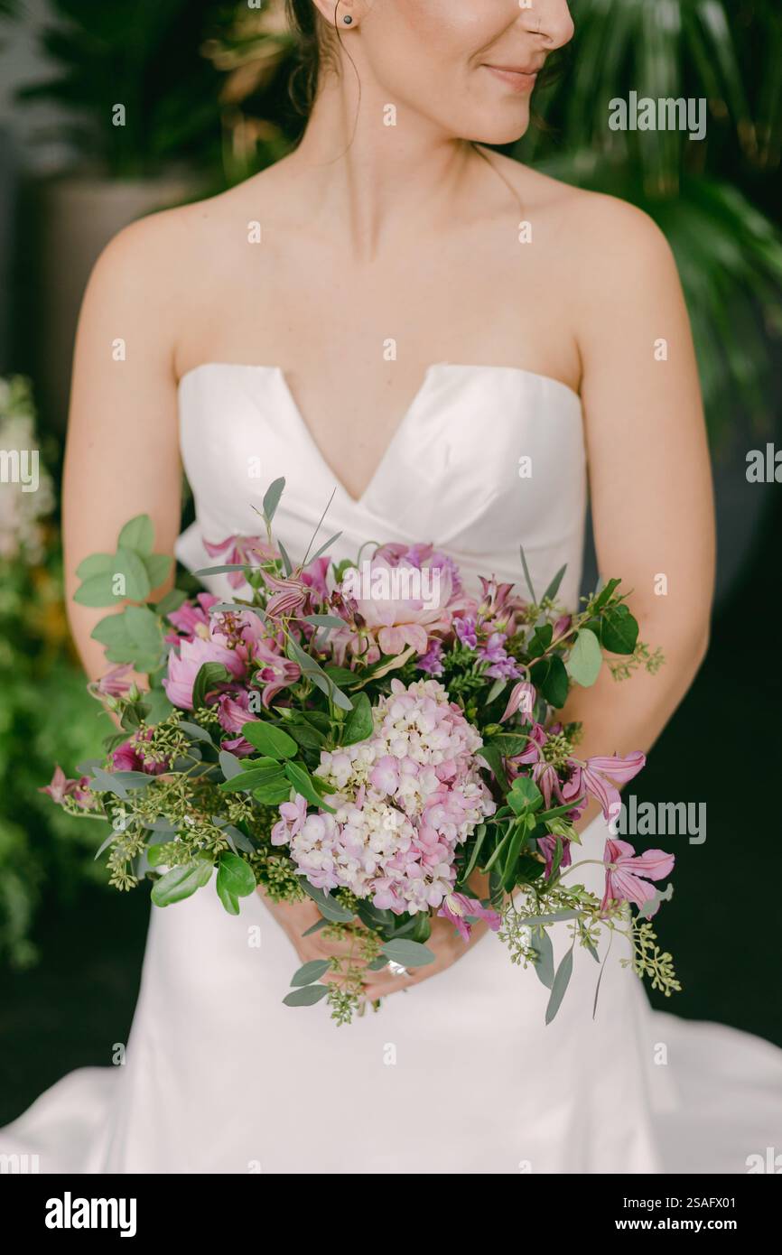 Una sposa mostra con orgoglio un bellissimo bouquet pieno di fiori viola e verdi mentre indossa un elegante abito senza spalline in una location per matrimoni Foto Stock