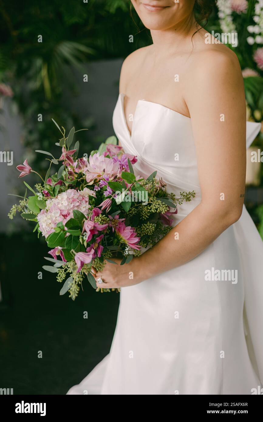 Una sposa sfoggia il suo elegante abito senza spalline mentre tiene in mano un bouquet colorato pieno di fiori viola e rosa in un giardino verde. L'atmosfera è ser Foto Stock