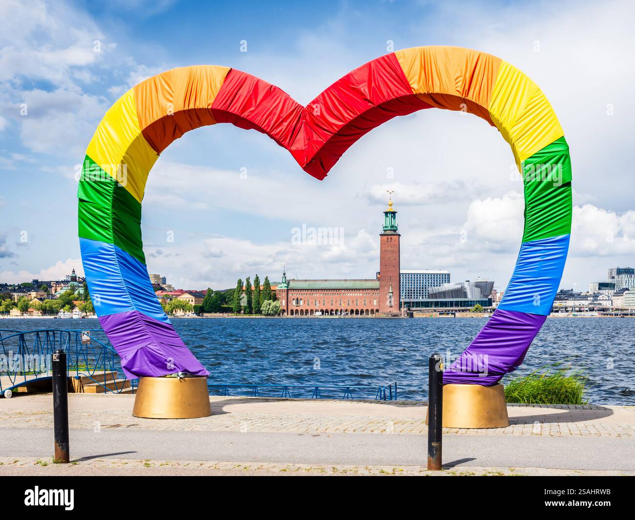 Il municipio si vede attraverso una struttura a forma di cuore ricoperta di tessuto nei colori della bandiera arcobaleno a Stoccolma, in Svezia, in una soleggiata giornata estiva. Foto Stock